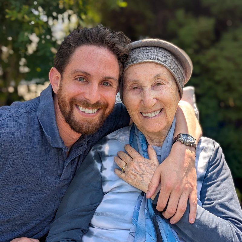 Moshe Birnbaum smiling with his arm around his grandmother, who rests her hand on her heart