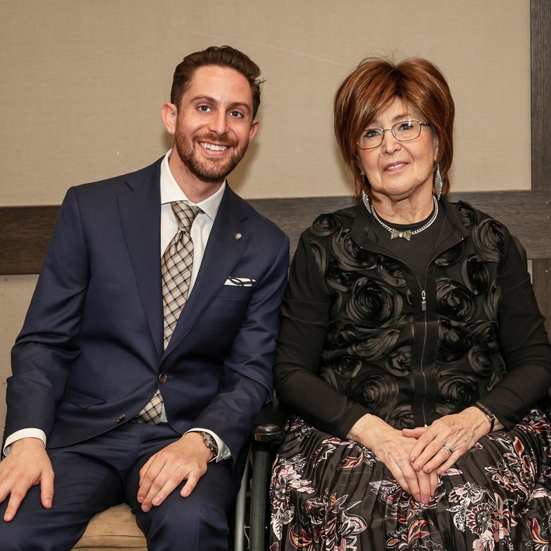 Moshe Birnbaum in a navy suit sitting beside his other grandmother, who is smiling in a dark floral outfit